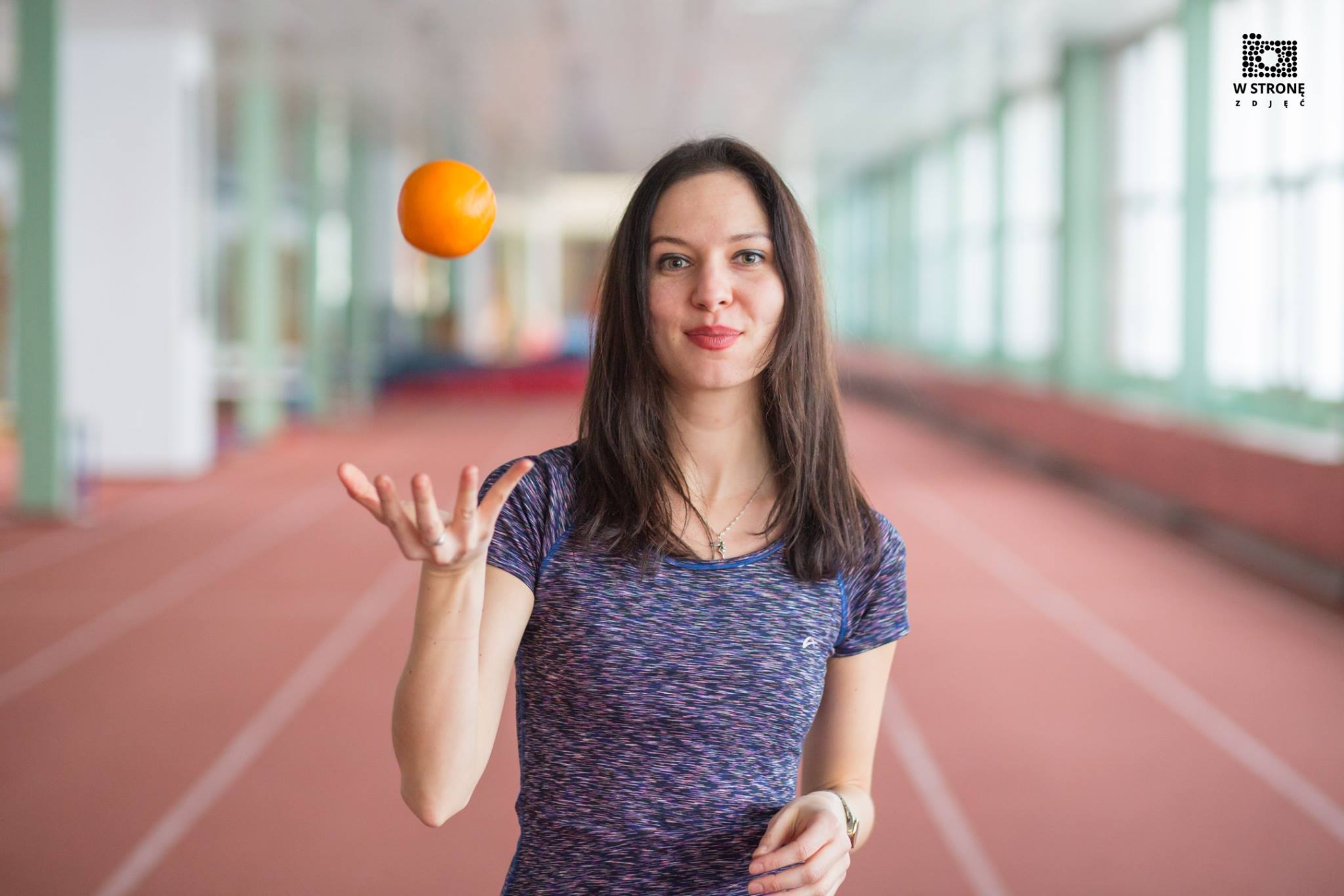 Maja Tomczyk - prowadząca badanie podrzuca pomarańczę - zdjęcie w fioletowym t-shircie na tle hali sportowej z bieżnią