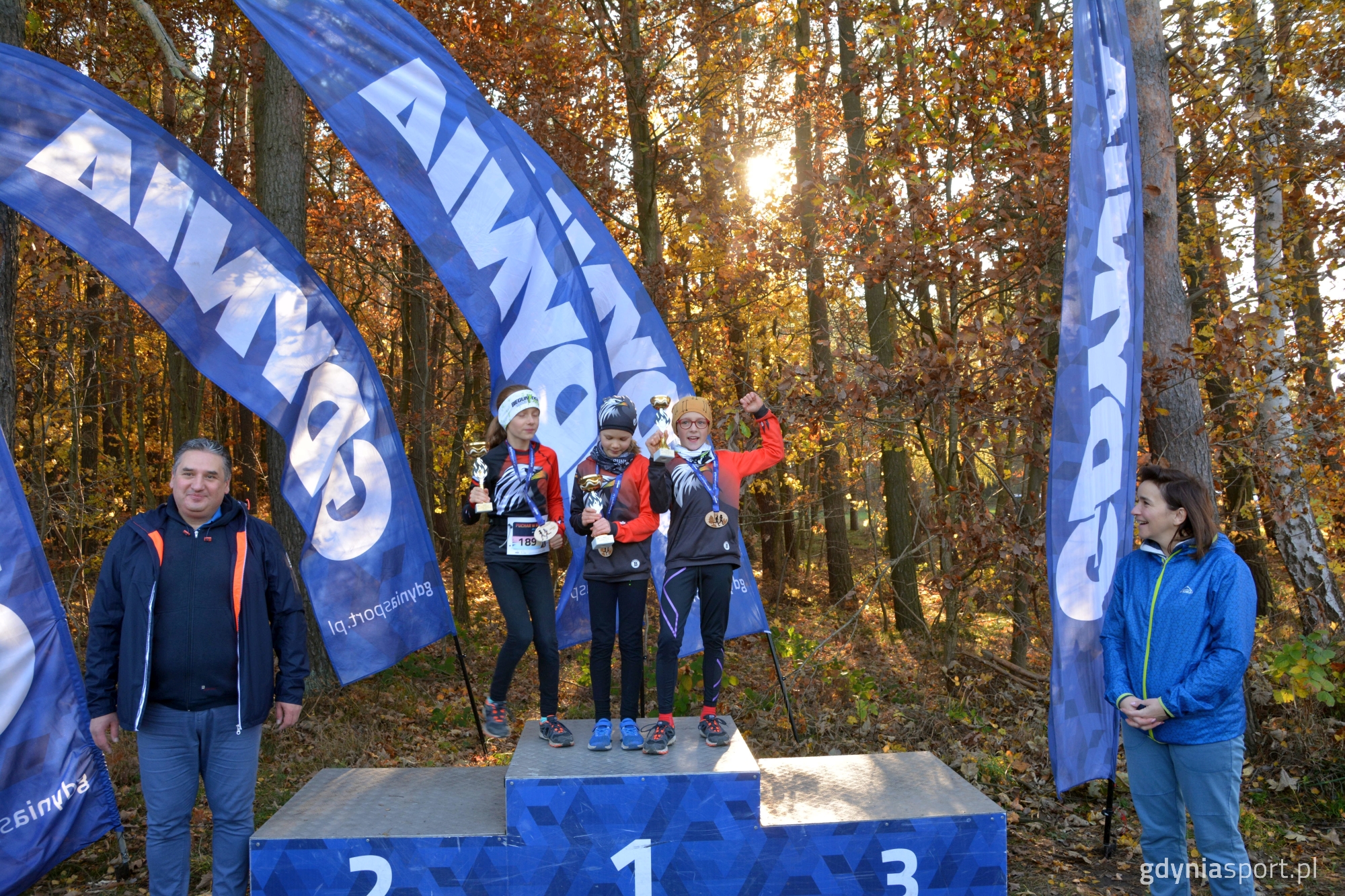 Najlepsze zawodniczki w sowjej kategorii w towarzystwie wręczających Anny Szpajer i Piotra Szpajer na tle lasu u słońca
