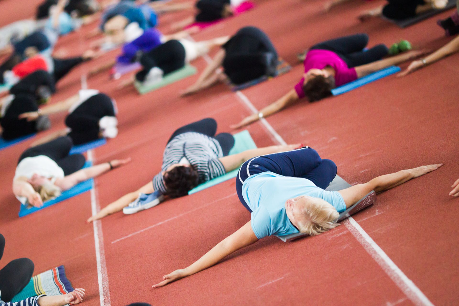 grupa ludzi leżących na ziemi ćwiczących pilates na hali lekkoatletycznej 