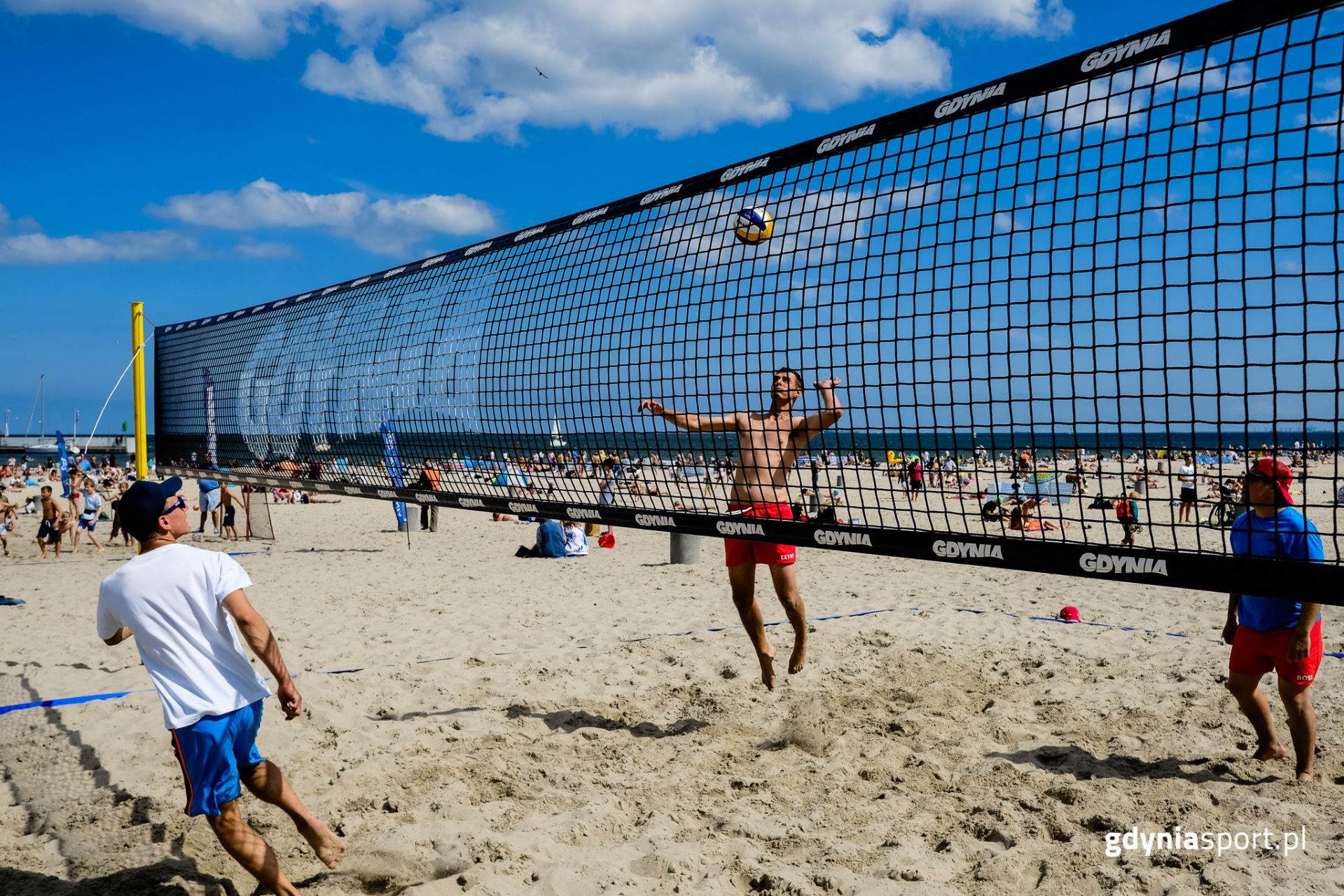chłopak w podskoku atakujący piłkę podczas gry w siatkówkę plażową. w tle plażowicze i morze 
