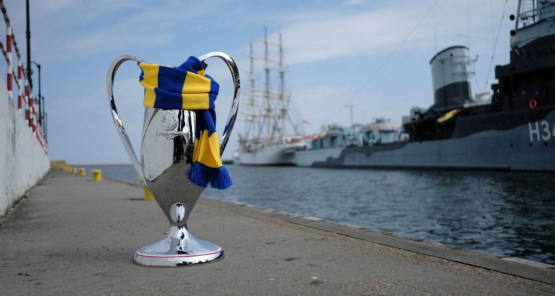 Puchar Polski owinięty żółto-niebieskim szalikiem stojący na jednym z nabrzeży. W tle ORP Błyskawica 