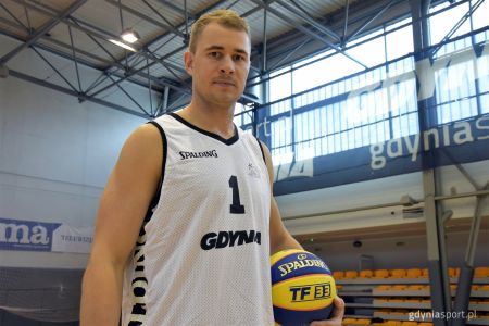 Bartłomiej Wołoszyn z w białej turniejowej koszulce stojący na hali gier gcs z żółto-niebieską piłką firmy Spalding